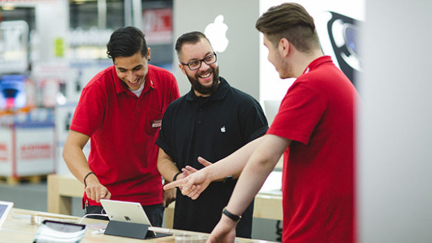 Drei Männer, einer mit Brille, lächeln und zeigen auf ein Tablet in einem Geschäft mit Apple Logo.