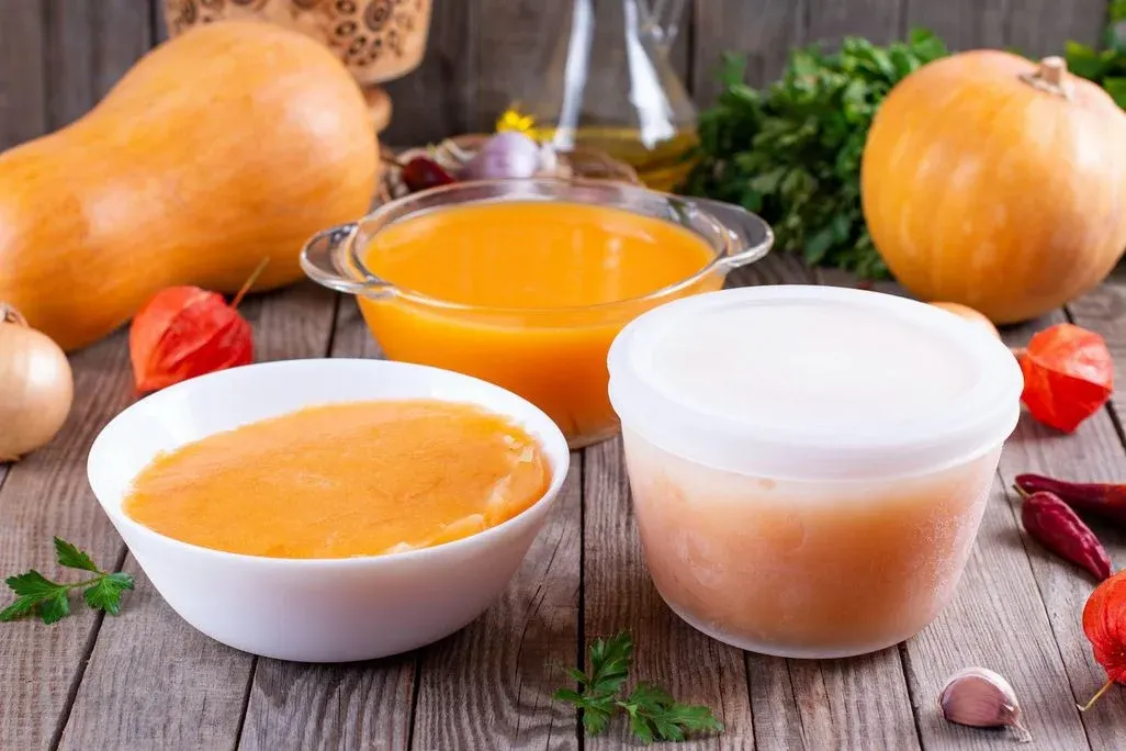 Drei Behälter mit Kürbispüree/-suppe, umgeben von Kürbissen, Zwiebeln, Knoblauch, Chilis und Physalis auf Holz.