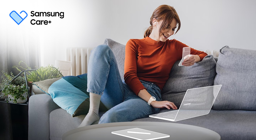 Eine Frau sitzt auf einem grauen Sofa und benutzt einen Laptop. Sie hält ein Smartphone in der Hand.