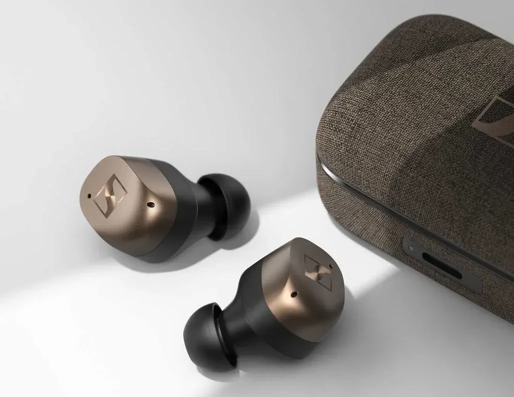 Ein Paar Sennheiser In-Ear-Kopfhörer in Schwarz und Kupfer, die neben ihrem geschlossenen, stoffbezogenen Ladecase liegen.