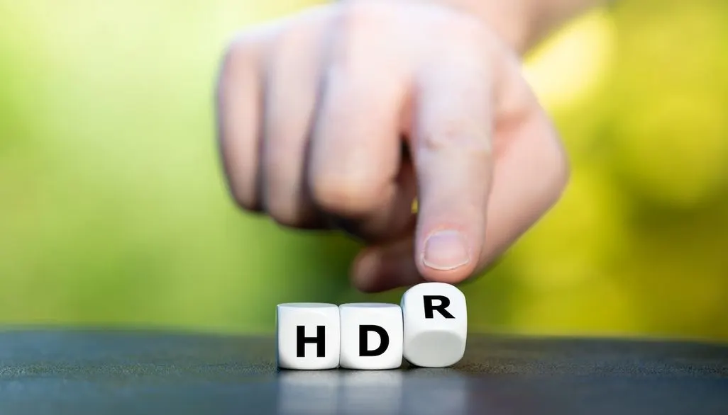 Eine Hand dreht einen Würfel mit der Aufschrift HDR auf einem Tisch, vor unscharfem grünen Hintergrund.