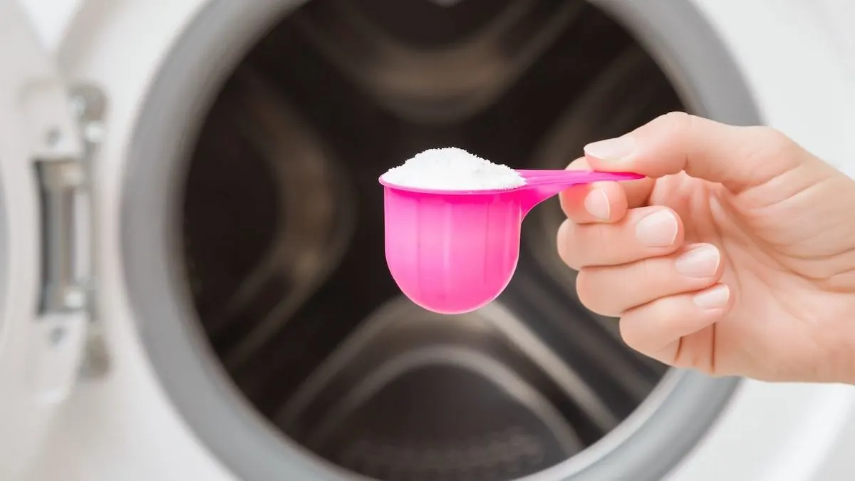 Eine Hand hält einen pinken Messbecher mit weißem Waschpulver vor einer geöffneten weißen Waschmaschine.