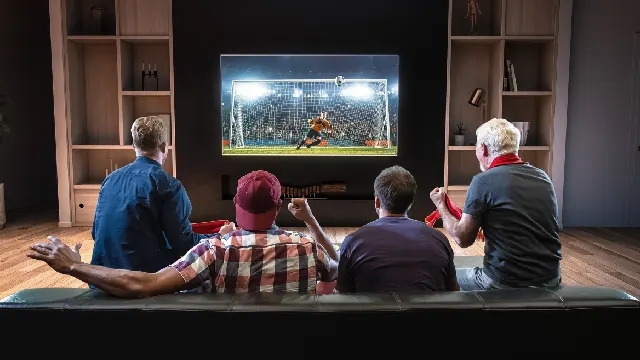 Vier Personen schauen Fußball im Fernsehen. Sie sitzen auf einer schwarzen Couch vor einem Bildschirm. Regale sind sichtbar.