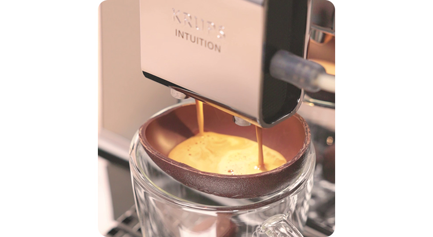  Krups Intuition Kaffeemaschine lässt Espresso in eine halbierte Schoko-Eierschale laufen, die in einem Glas steht – kreative Osterkaffee-Idee.