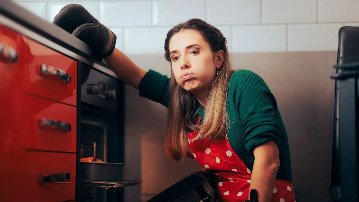 Person mit roter Punkteschuerze und Ofenhandschuh nimmt einen Kuchen aus dem Backofen in einer modernen Küche.