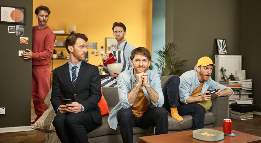 Eine Gruppe von Männern in einem Wohnzimmer, einige sitzen auf einer Couch, einer spielt ein Videospiel, ein anderer schaltet das Licht aus.