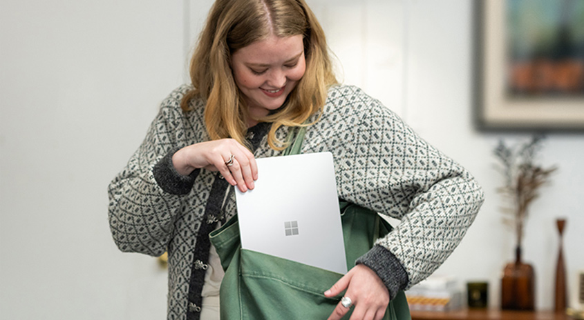 Eine Frau legt einen silbernen Laptop in eine grüne Tasche. Sie trägt einen gemusterten grauen Pullover. Im Hintergrund ist eine Wand und ein Tisch.