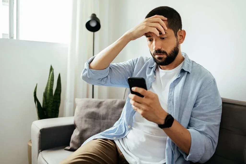 Ein Mann auf einer grauen Couch blickt besorgt auf sein schwarzes Smartphone, die Hand an der Stirn. Im Hintergrund sind eine grüne Pflanze und eine schwarze Stehlampe zu sehen.