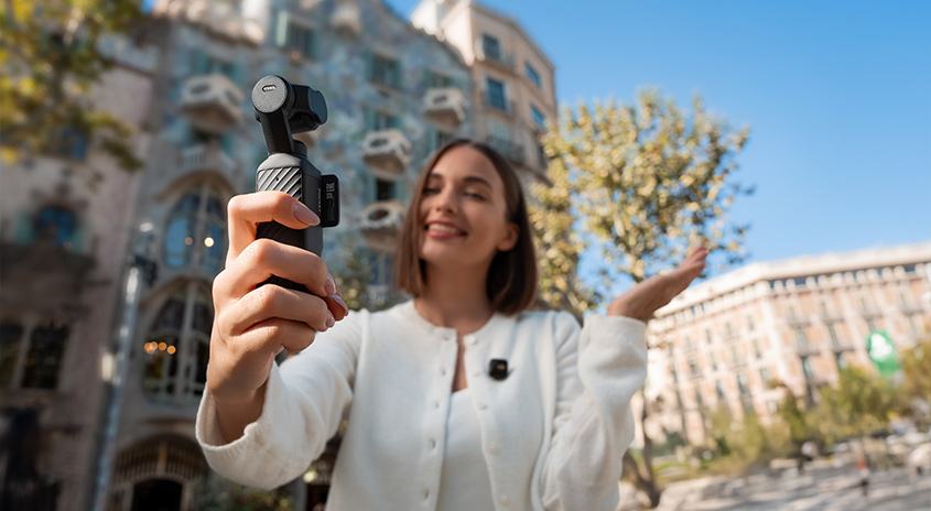 Frau in weißer Jacke hält DJI Osmo Pocket Kamera-Gimbal in die Höhe. Im Hintergrund Architektur einer europäischen Stadt mit blauem Himmel.