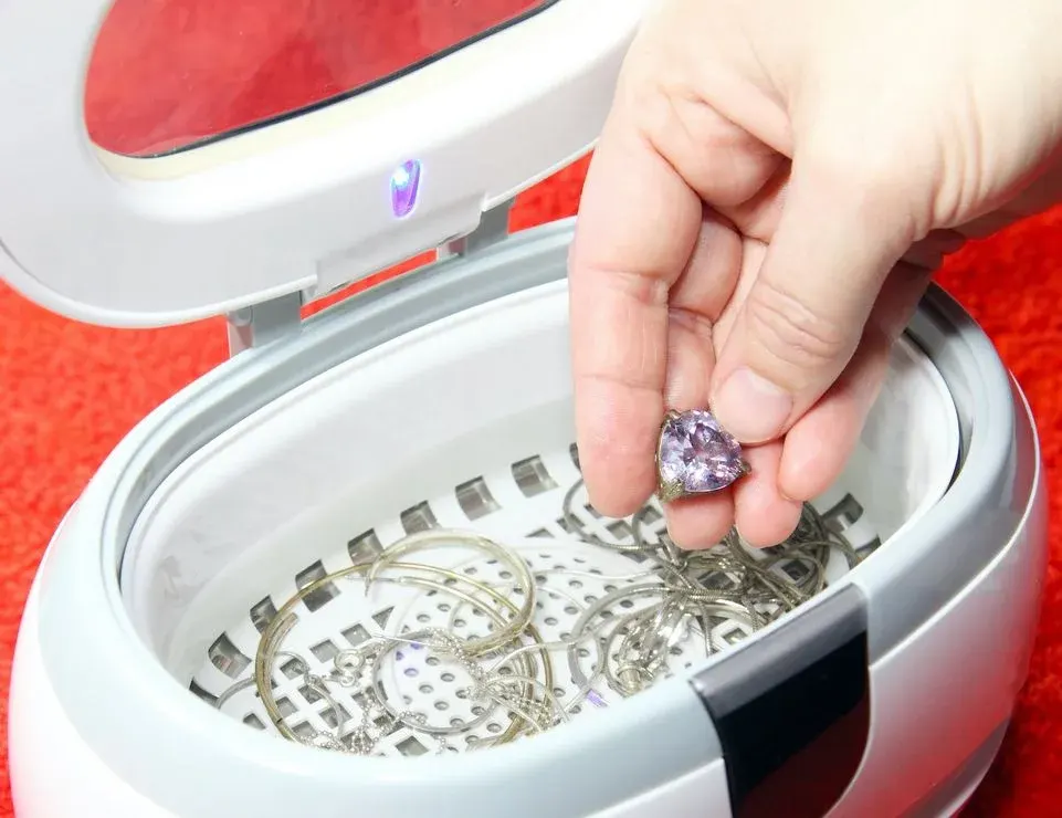 Hand hält lila Herz-Anhänger über geöffnetem weißem Ultraschallreiniger. Silberner Schmuck liegt in Wasser auf rotem Hintergrund.