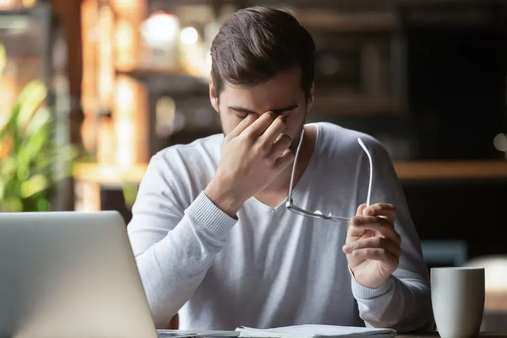 Man sitzt vor dem Laptop und eine Kaffeetasse steht am Schreibtisch. Er hält die Brille mit der linken Hand und reibt sich die Augen.