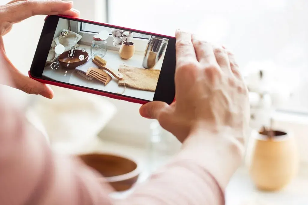Hände halten ein rotes Smartphone horizontal, das eine Zusammenstellung aus Holzschale, Bürste, Kamm, Glas, Metallbecher, Stoffbeutel und Baumwollblüten zeigt.
