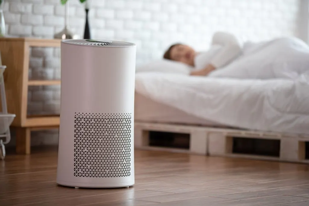 Luftreiniger steht in einem modernen Schlafzimmer, im Hintergrund ist eine schlafende Person im Bett zu sehen.