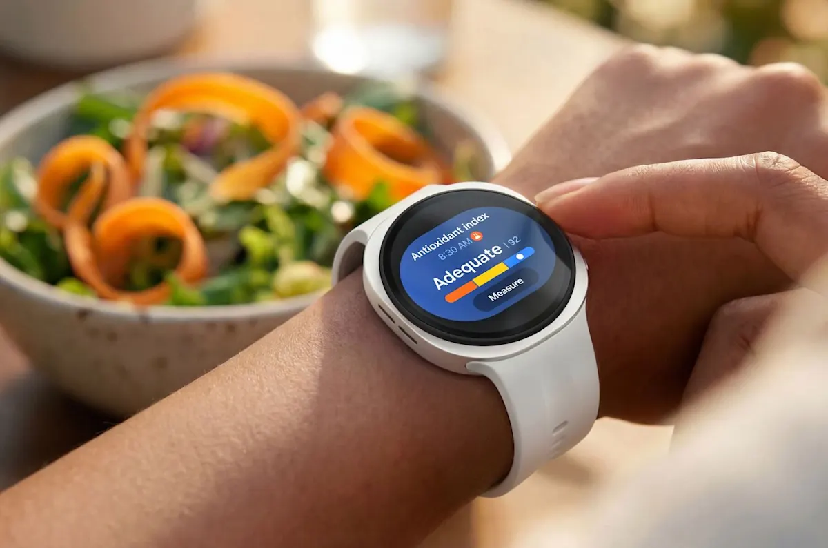 Eine Samsung Galaxy Watch9 mit weißem Armband auf einem Handgelenk. Auf dem Display wird der Antioxidant-Index angezeigt. Im Hintergrund steht eine Schüssel Salat
