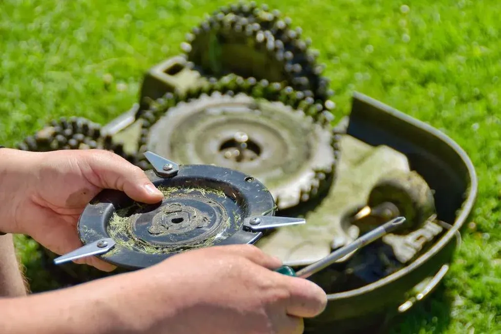 Hände wechseln das Messerteller mit Schneidklingen an der Unterseite eines geöffneten Rasenmähroboters auf einer Wiese.