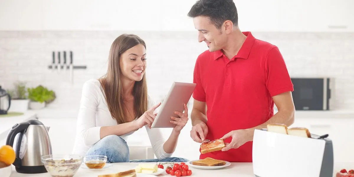 Paar in weißer Küche beim Frühstück: Frau mit Tablet, Mann streicht Marmelade auf Toast. Wasserkocher, Müsli, Toaster auf dem Tresen.