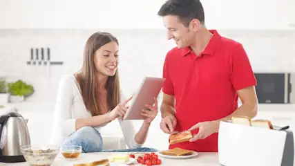Paar in weißer Küche beim Frühstück: Frau mit Tablet, Mann streicht Marmelade auf Toast. Wasserkocher, Müsli, Toaster auf dem Tresen.