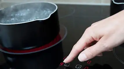 Hand bedient ein Touchfeld am Kochfeld neben einem Topf kochenden Wassers