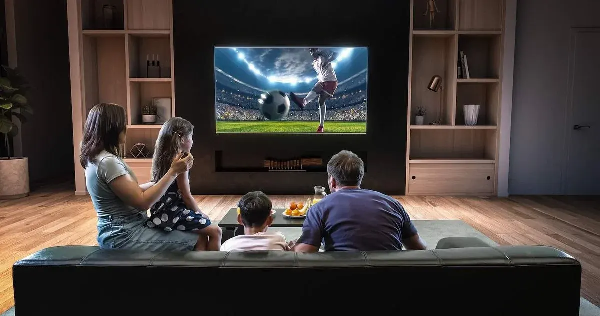 Familie sitzt in einem abgedunkelten Wohnzimmer auf der Couch und schaut Fußball am Fernseher.