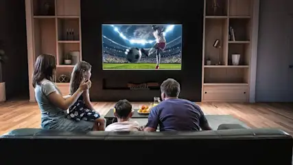 Familie sitzt in einem abgedunkelten Wohnzimmer auf der Couch und schaut Fußball am Fernseher.