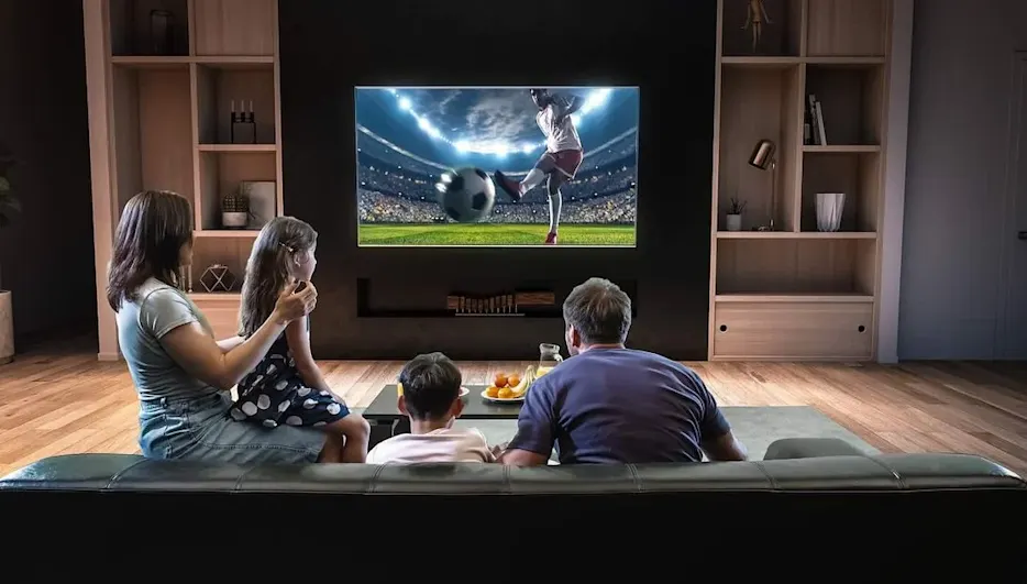 Familie sitzt in einem abgedunkelten Wohnzimmer auf der Couch und schaut Fußball am Fernseher.