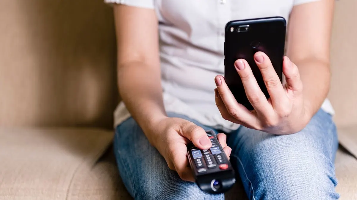 Person in weißem Hemd und blauen Jeans hält schwarzes Smartphone und schwarze TV-Fernbedienung auf beigem Sofa.