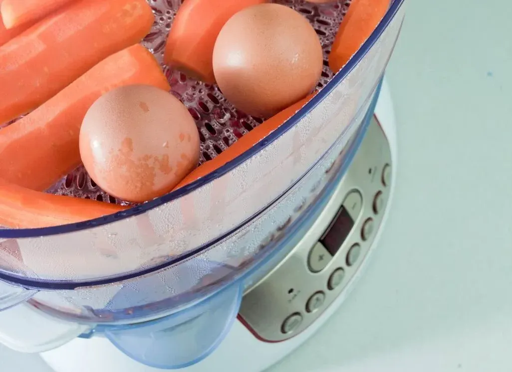 Elektrischer Dampfgarer mit transparentem Behälter, in dem Karottenstücke und braune Eier auf einem gelochten Einsatz gleichzeitig schonend gegart werden.