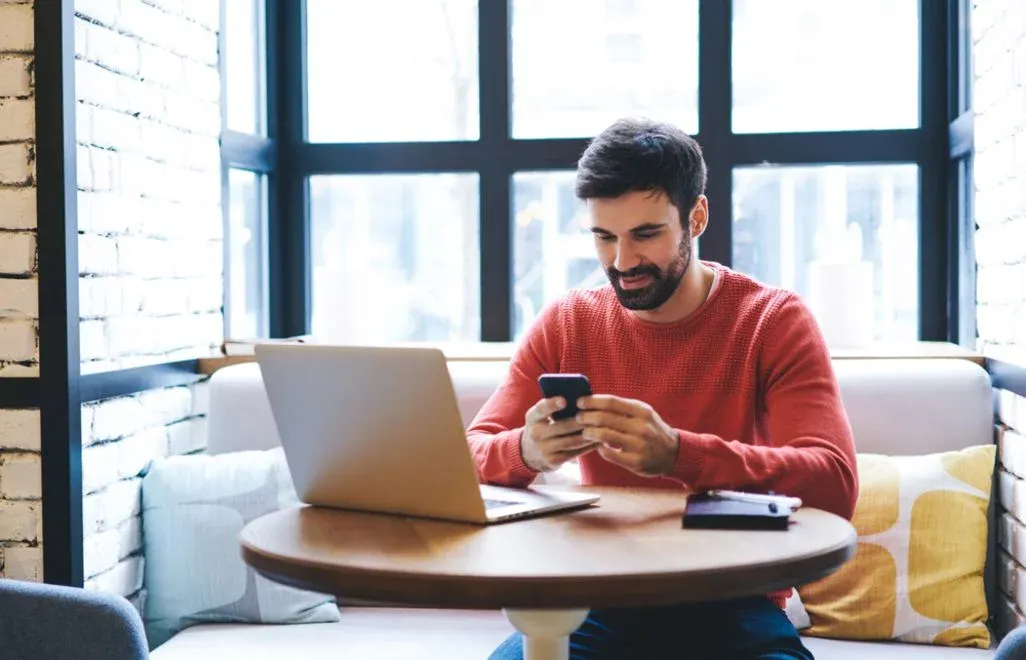 Ein Mann in rotem Pullover sitzt an einem Holztisch, blickt auf sein Smartphone. Ein Laptop und eine Geldbörse liegen auf dem Tisch.