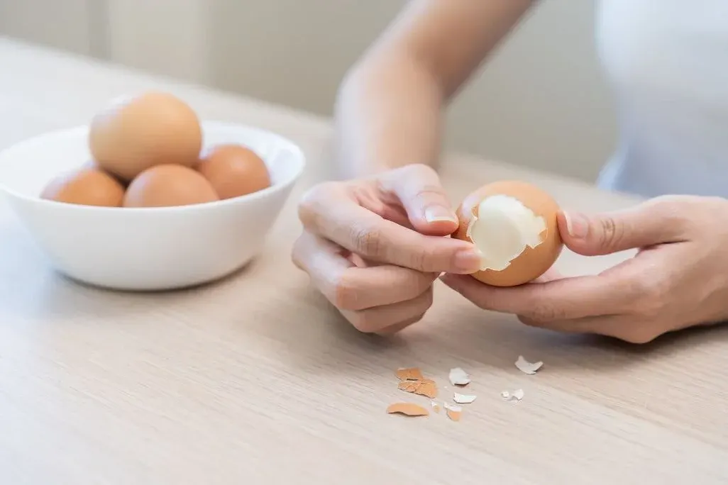 Person schält ein hart gekochtes Ei über einem Tisch, Eierschalen liegen daneben, im Hintergrund steht eine weiße Schale mit mehreren braunen Eiern.