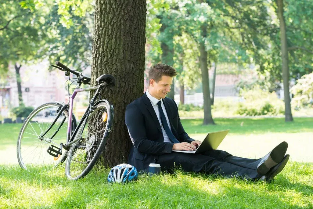Ein Mann im Anzug sitzt lächelnd unter einem Baum, arbeitet am Laptop. Ein Fahrrad, Helm und Kaffeebecher sind daneben auf grünem Gras.