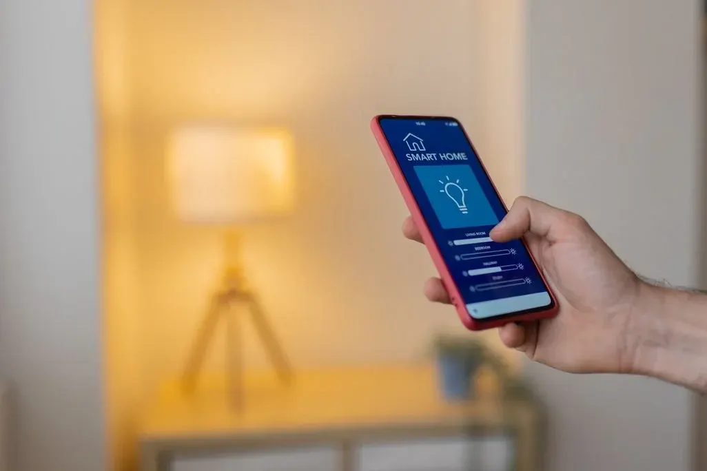 Eine männliche Hand hält ein Smartphone mit rotem Gehäuse. Auf dem Display ist eine Smart-Home-App mit blauem Hintergrund geöffnet. Die App zeigt das Icon eines Hauses, die Aufschrift „Smart Home" sowie eine leuchtende Glühbirne und Schieberegler zur Helligkeitssteuerung für mehrere Räume (Living Room, Bedroom, Hallway, Study). Im unscharfen Hintergrund ist ein Wohnraum mit einer warm leuchtenden Stehlampe auf einem Holzstativ zu sehen.