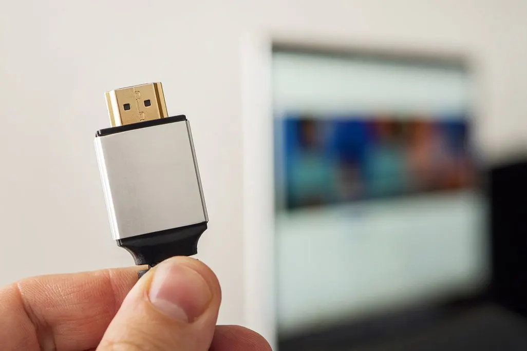 Hand hält einen HDMI-Stecker vor einen verschwommenen Bildschirm.