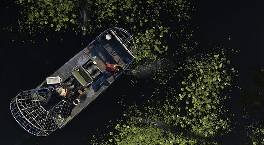Zwei Leute in Grassrivers in einem Sumpfboot aus der Vogelperspektive, umgeben von Alligatoren. Ein Mann zielt mit einer Waffe auf den Alligator, der ihm am nächsten ist.