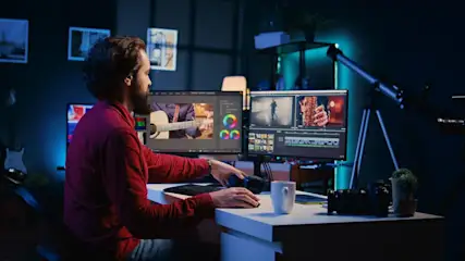 Bärtiger Mann in rotem Hemd bearbeitet Videos an zwei Monitoren mit Bearbeitungssoftware. Auf dem Tisch Kamera, Tasse. Im dunklen Raum Gitarre, Teleskop.