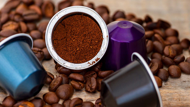 Kaffeebohnen und Kapseln in Blau, Lila und Schwarz liegen auf einem Holztisch. Eine Kapsel ist offen und mit Kaffeepulver gefüllt.