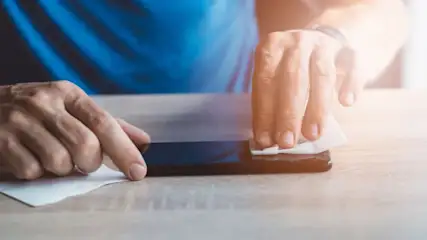 Hände reinigen ein schwarzes Smartphone mit einem weißen Tuch auf einem hellen Holztisch; blauer Stoff im Hintergrund.