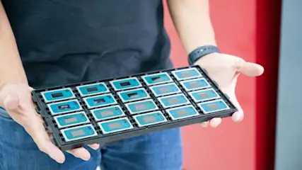 Eine Person hält ein schwarzes Tablett mit 18 rechteckigen Chips mit blauen und grauen Elementen vor rotem und grauem Hintergrund.