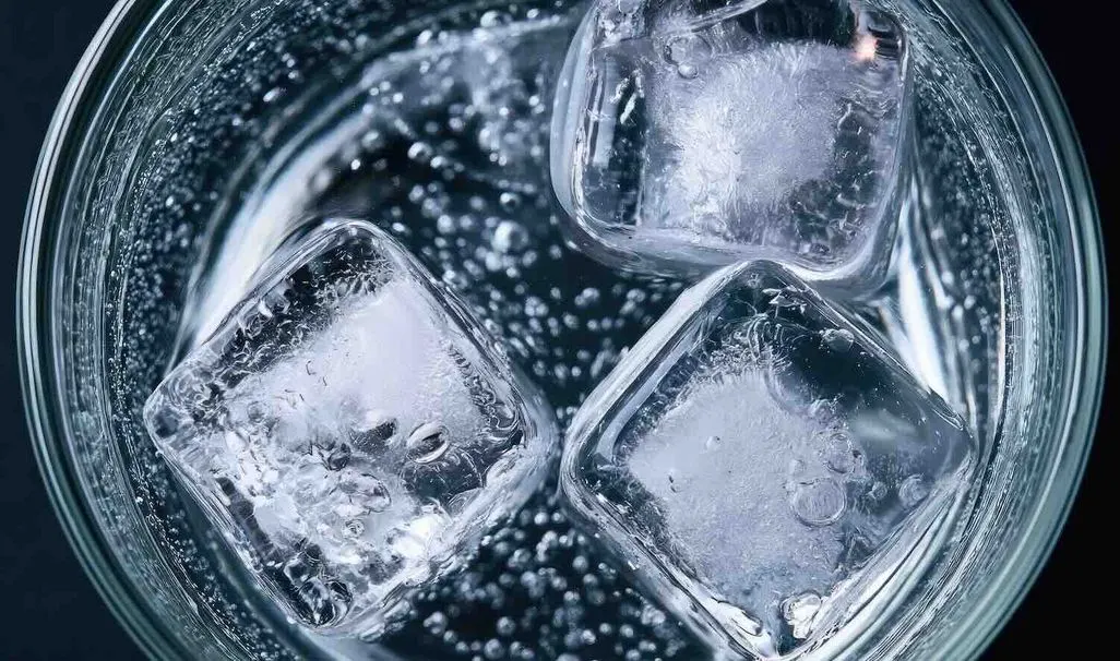 Draufsicht in ein Glas mit drei Eiswürfeln und sprudelndem Wasser, kleine Luftbläschen steigen auf.