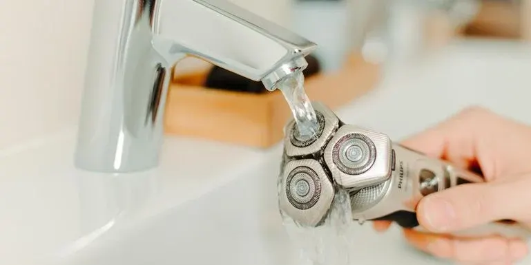 Eine Hand hält einen silbernen Elektrorasierer unter fließendem Wasser aus einem glänzenden Wasserhahn.