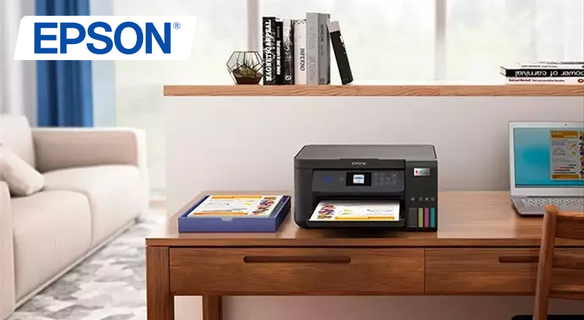 Ein grauer Drucker steht auf einem Holztisch, neben einem Laptop und einer Ablage. Bücher und Deko stehen auf Regalen im Hintergrund.