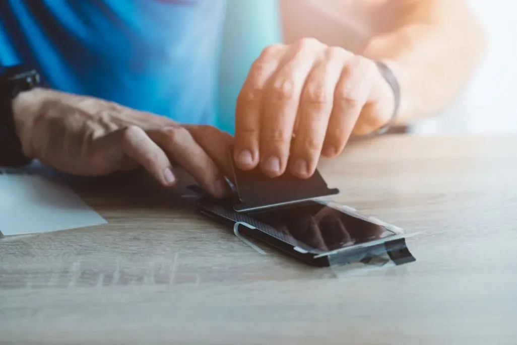 Person klebt eine Schutzfolie auf das Display eines Smartphones, das auf einem Holztisch liegt.