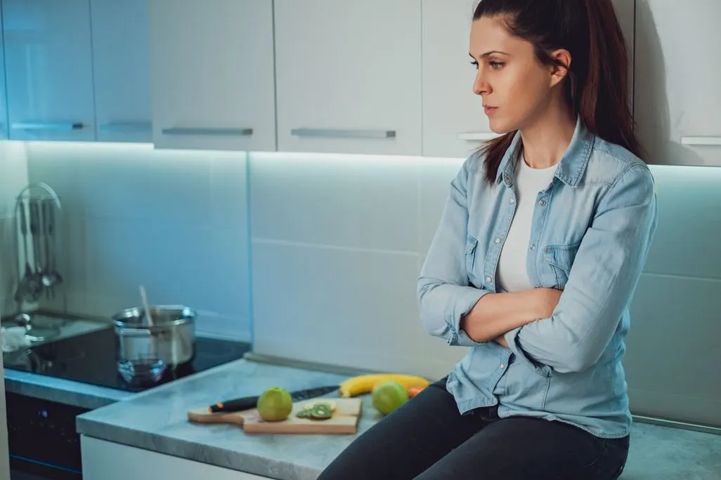Frau sitzt mit verschraenkten Armen auf Kuechentheke neben geschnittenem Obst