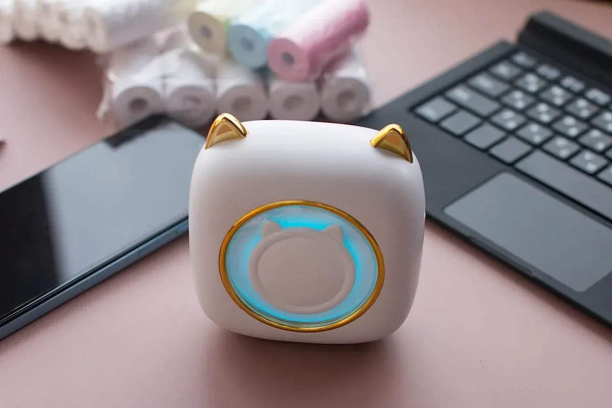 Weißes Katzen-Gadget mit goldenem Ohren und blauem Kreis-Display auf rosa Schreibtisch neben schwarzer Tastatur und Rollen von Klebeband – technisches Zubehör für Büro oder Heimarbeitsplatz.