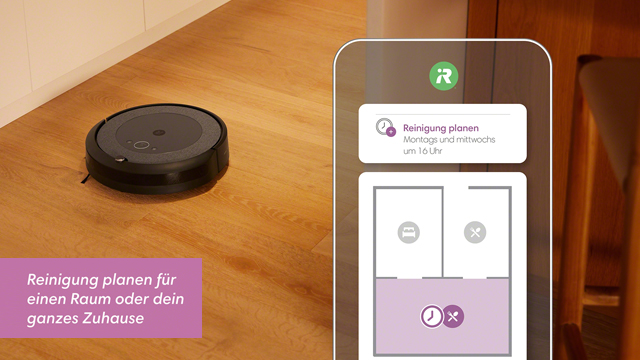 Ein schwarzer Saugroboter steht auf einem Holztisch. Daneben ist ein Smartphone mit einer Reinigungsplan-App zu sehen.