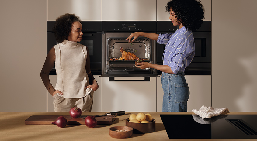 Eine Küche mit einem Ofen, in dem ein Braten zubereitet wird. Auf der Arbeitsplatte liegen Zwiebeln, Äpfel, Kartoffeln und ein Messer.