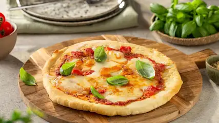 Eine Pizza mit Tomatensoße, Mozzarella und Basilikum liegt auf einem Holzbrett. Im Hintergrund sind Tomaten, frischer Basilikum und Gewürze zu sehen.