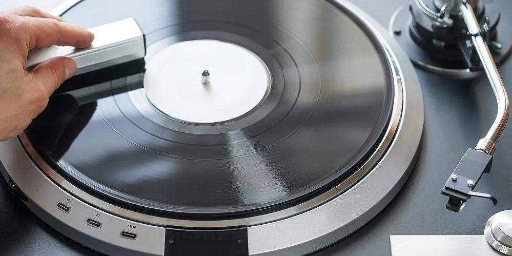 Eine Hand reinigt eine schwarze Vinyl-Schallplatte mit einer silbernen Bürste auf einem silbernen Plattenspieler. Weiße Etikette und Tonarm sichtbar.