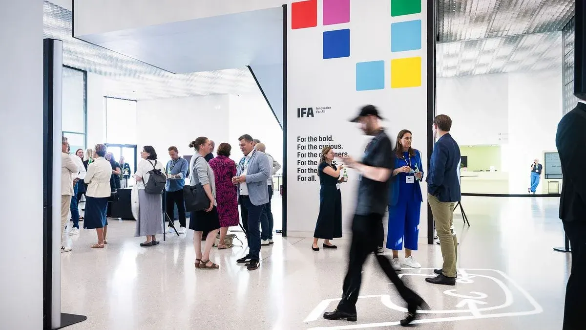 Personen unterhalten sich in einer modernen Halle. Eine weiße Wand zeigt farbige Quadrate und IFA-Logo. Einige gehen, andere stehen mit Getränken. Heller Schalter im Hintergrund.