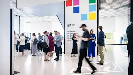 Personen unterhalten sich in einer modernen Halle. Eine weiße Wand zeigt farbige Quadrate und IFA-Logo. Einige gehen, andere stehen mit Getränken. Heller Schalter im Hintergrund.