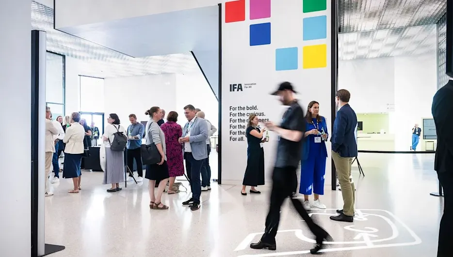 Personen unterhalten sich in einer modernen Halle. Eine weiße Wand zeigt farbige Quadrate und IFA-Logo. Einige gehen, andere stehen mit Getränken. Heller Schalter im Hintergrund.